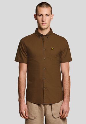 Jeune homme aux cheveux courts portant une chemise marron à manches courtes avec un logo d'oiseau jaune sur la poche poitrine et un pantalon beige.