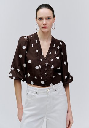 Frau trägt eine braune, kurz geschnittene Bluse mit weißen Polka-Dots und Puffärmeln, kombiniert mit hoch taillierten weißen Hosen und Creolen.