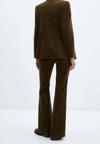 Ruskea maneesimuotoinen puku, jossa on istuva bleiseri ja levenevät housut, napin koristuksilla ja sileällä tekstuurilla.