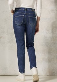 Vaqueros de mezclilla azul oscuro con un corte ajustado, que cuentan con bolsillos traseros y costuras sutiles. Combinados con zapatillas deportivas blancas de caña alta.