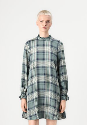 Vihreä ruudullinen mekko, jossa on röyhelökaulus ja pitkät hihat. Kangas sisältää tekstuuripintaisen kuosin, jossa on valkoisia ja tummansinisia yksityiskohtia.