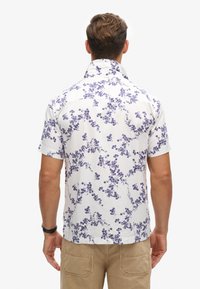Camisa blanca de manga corta con patrones florales morados, diseño con cuello y dobladillo ligeramente curvado, mostrada desde la parte posterior.