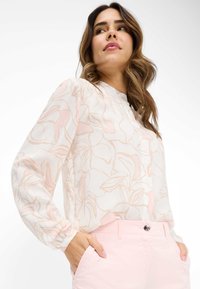 Blusa bianca con motivo floreale rosa chiaro, maniche lunghe e scollatura tonda. Modella indossa pantaloni rosa chiaro, con la mano in tasca.