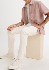 Vaaleanpunainen lyhythihainen pellavapaita taskulla, yhdistetty kermaisiin farkkuihin ja vaaleanbeigeihin korkean varren lenkkitossuihin puisella korokkeella.