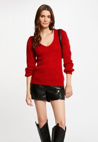Maglione rosso lavorato a maglia con scollatura a V, maniche a sbuffo lunghe e texture a coste, abbinato a pantaloni corti in pelle nera e stivali alti neri.