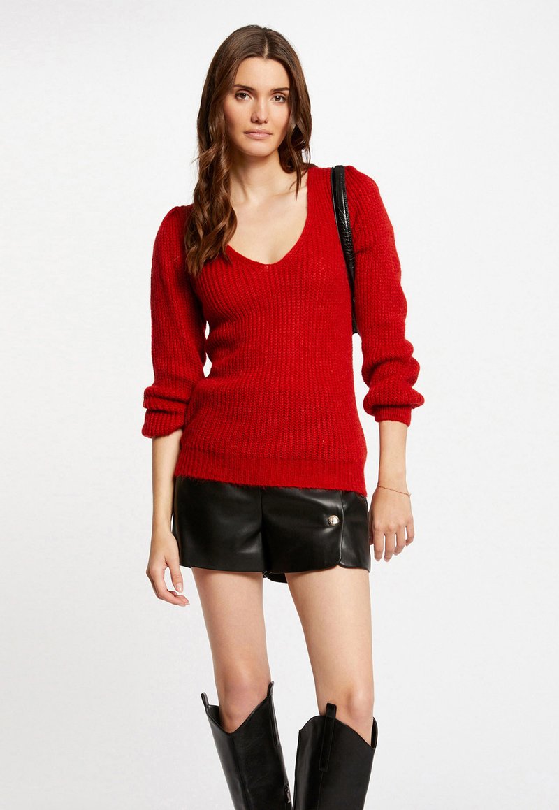 Maglione rosso lavorato a maglia con scollatura a V, maniche a sbuffo lunghe e texture a coste, abbinato a pantaloni corti in pelle nera e stivali alti neri.