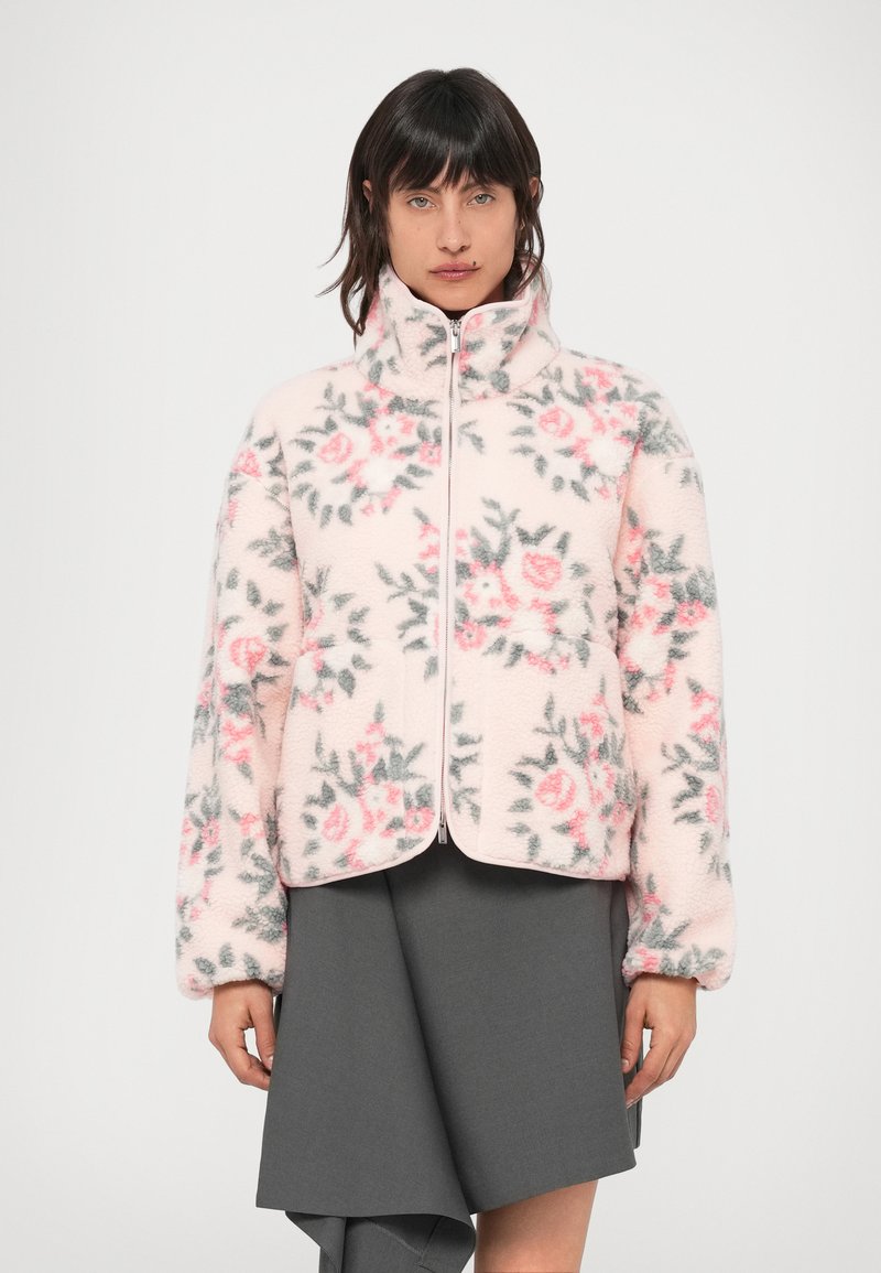Fleecová bunda v jemne ružovej farbe s kvetinovými vzormi v ružovej a šedej. Má vysoký golier a zapínanie na zips, zaoblené lemy a nadýchané rukávy.