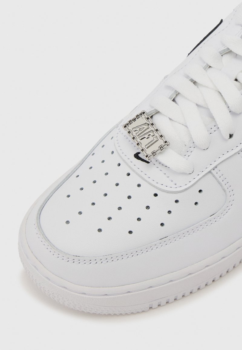 Bout de basket blanche avec un design perforé, des lacets blancs et une étiquette métallique gravée "AF1" sur un fond uni.