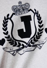 Pelēka T-krekls ar melnu vairogu logotipu, kas rotāts ar kroni un lauru lapām, kurā izceltas diženas burti "J" un teksts "Juicy Couture" un "League."