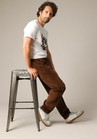 Pantalons en velours côtelé marron et T-shirt graphique blanc avec un design. Le mannequin est assis sur un tabouret en argent portant des baskets blanches avec des semelles marron.