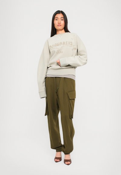 Young woman wearing beige DSQUARED2 sweatshirt, olive green cargo pants, and brown open-toe heels, standing against a plain white background.