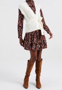 Gilet en tricot blanc avec une ceinture à nouer sur une robe maroon à motif paisley. Des bottes en suède marron jusqu'aux genoux complètent le look.