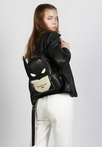 Mochila de cuero negro con un diseño tridimensional de Batman que incluye orejas, ojos blancos y un panel de cara beige, asegurada con una cremallera.