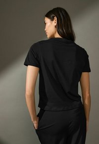 T-shirt nera a maniche corte con scollo rotondo e vestibilità morbida, realizzata in tessuto liscio, indossata con pantaloni neri.