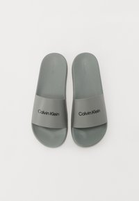 Sandale gri cu talpă joasă, cu un finisaj mat, având o curea largă imprimată cu "Calvin Klein" în negru. Tălpică netedă și moale.