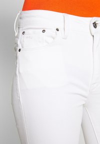 Vaqueros de denim blanco con una textura suave, diseño de cinco bolsillos y detalles metálicos. Presenta un sutil logotipo de la marca en el bolsillo trasero.