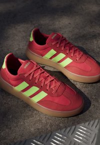 Pair of red Adidas sneakers with neon green stripes and gum soles placed on a dark textured surface in soft light.