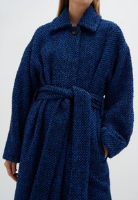 Texturerad blå kappa med herringbone-mönster, överdimensionerad krage, framknäppning med knappar och bälte i midjan. Långa ärmar täcker händerna.