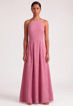 Donna che indossa un vestito rosa senza maniche, con collo halter e lunghezza fino a terra, in piedi davanti a uno sfondo chiaro e uniforme.