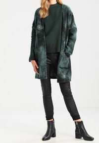 Cardigan lungo verde morbido con un motivo testurizzato, indossato sopra un maglione verde, abbinato a pantaloni neri attillati e stivaletti.