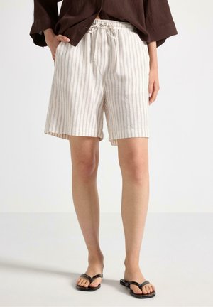 Person iført beige og hvide lodrette stribede shorts med snøre og lommer, mørkebrun top og sorte thongsandaler, stående.