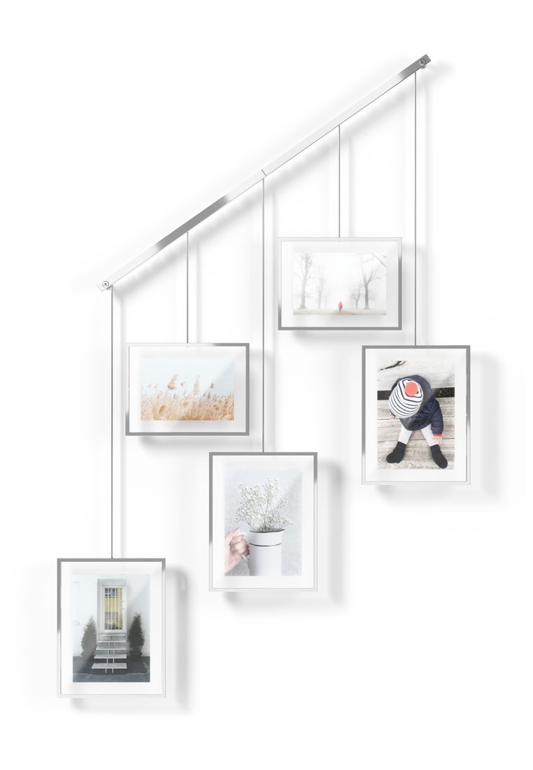 Exhibición de fotos montada en la pared que presenta cinco marcos rectangulares de vidrio transparente con bordes plateados, suspendidos por finos cables de una barra metálica en ángulo.
