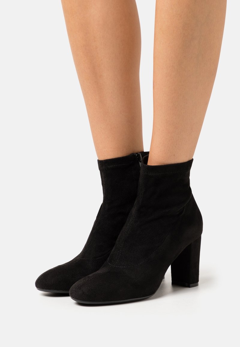 Femme portant des bottines en daim noir à talons blocs sur un fond clair uni.
