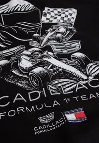 Cadillac Formel 1 racerbiler, sjakkmønstrete flagg og logoer for Cadillac og Tommy Hilfiger på svart stoffbakgrunn.
