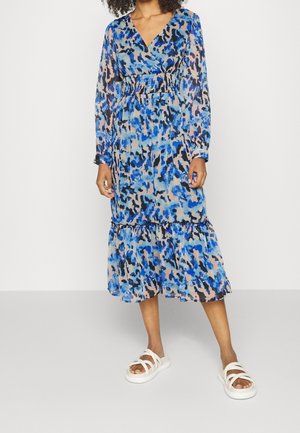 Robes à imprimé animal bleu et beige avec des manches longues, taille smockée et ourlet à volants. Portée avec des sandales blanches à lanières.