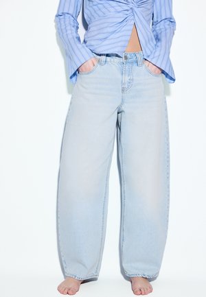 Osoba oblečená v svetlomodrých širokých džínsoch a modrej pásikavej dlhorukávovej košeli, stojaca bosá s rukami v vreckách.