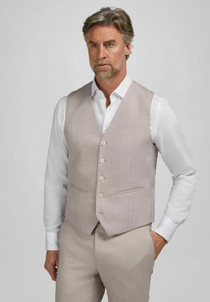 Mann trägt eine beige Anzugweste und Hose mit einem weißen Hemd, steht mit einer Hand in der Tasche vor einem einfarbigen Hintergrund.