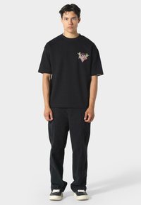 Musta puuvillane T-särk lühikeste varrukatega, millel on vasakul rinnal väike värviline tikand. Kantud koos sobivate mustade pükstega.