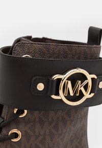 Hnědá vzorovaná bota s černým koženým popruhem, který má zlatý kulatý sponu s logem. Obsahuje očko na šněrování a texturovaný povrch.