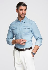 Chemise en jean bleu clair avec col pointu, deux poches poitrine, manches retroussées, associée à un pantalon blanc et une ceinture marron.