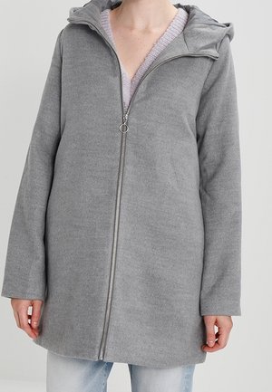 Femme portant un manteau gris à capuche mi-long zippé sur un pull lavande et un jean clair.