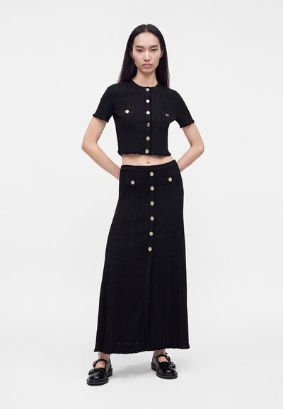 Femme aux longs cheveux noirs debout, portant un haut court noir à manches courtes et une longue jupe avec des boutons dorés, associée à des chaussures noires sur fond blanc.