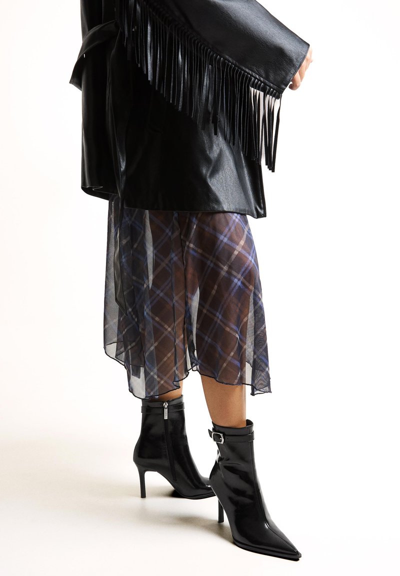 Femeie purtând o jachetă neagră din piele cu franjuri, fustă transparentă în carouri și cizme negre cu toc mediu, cu fermoar lateral și catarame.
