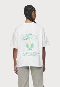 Weißes Baumwoll-T-Shirt mit grünem und gelbem grafischem Text auf dem Rücken, mit einem Sonnen- und Palmblattdesign. Lässige Passform und kurze Ärmel.