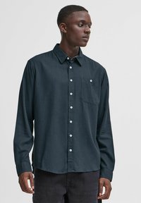 Camicia a maniche lunghe in tessuto blu scuro con chiusura a bottoni, colletto e una tasca sul petto. Presenta bottoni bianchi e una superficie testurizzata.