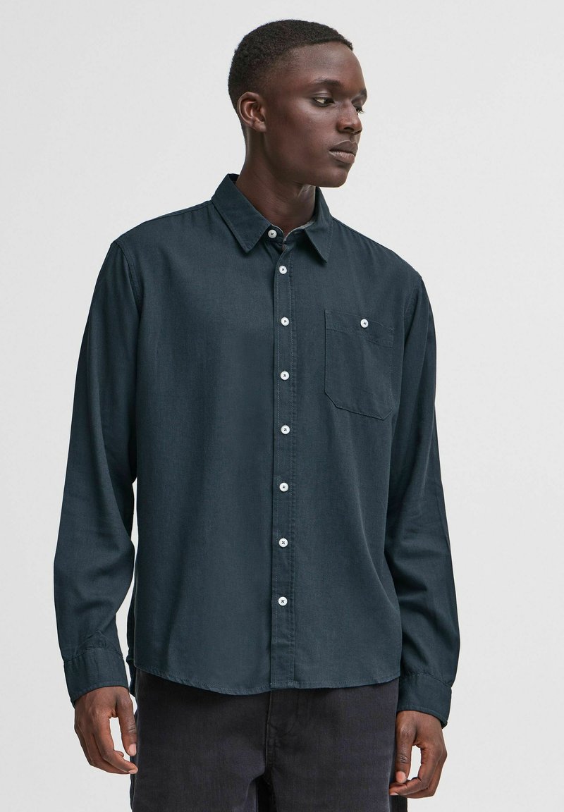 Camicia a maniche lunghe in tessuto blu scuro con chiusura a bottoni, colletto e una tasca sul petto. Presenta bottoni bianchi e una superficie testurizzata.