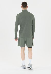Olijfgroene sporttop met lange mouwen en een hoge kraag, gecombineerd met olijfgroene shorts. Accessoires zijn grijze sneakers en witte sokken.