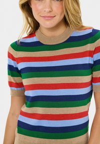 Vrouw in een kortlopende gebreide top met horizontale strepen in beige, rood, blauw, groen en marineblauw, die lichtjes glimlacht tegen een witte achtergrond.