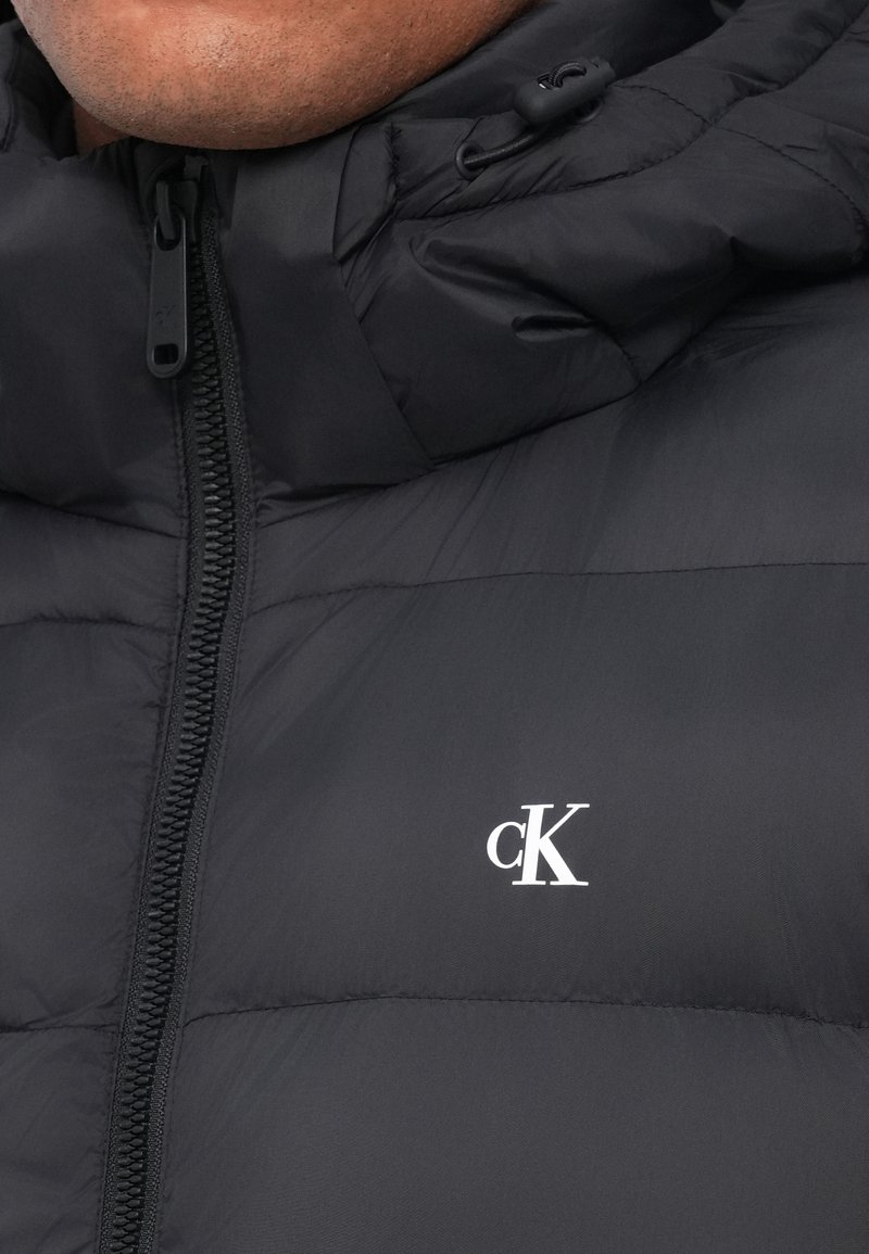 Schwarze Pufferjacke mit Reißverschluss, verstellbarem Kapuzenbereich und weißem "cK"-Logo auf der Brust. Glatte Textur und gestepptes Design.