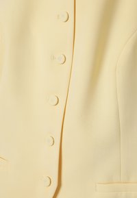 Gros plan sur le devant d'une veste jaune pâle avec cinq boutons recouverts de tissu et un détail subtil de poche latérale.