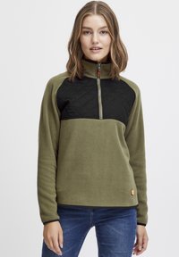 Pullover in pile di colore verde oliva con una sezione trapuntata nera sul petto. Presenta un colletto con zip a metà e un piccolo logo arancione sull'orlo.