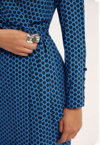 Niebieska sukienka w kształcie owijanej z czarnym geometrycznym wzorem, z paskiem w talii, długimi rękawami oraz srebrnym pierścionkiem na prawej dłoni.