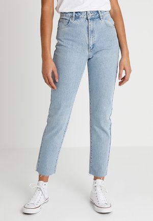 Osoba ubrana w jasnoniebieskie jeansy z wysokim stanem, białe trampki i białą koszulę, stojąca z rozluźnionymi ramionami na jednolitym tle.