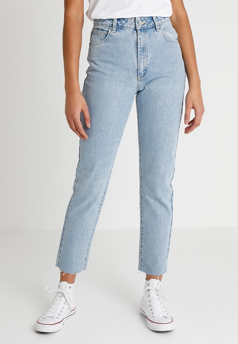 Personne portant un jean taille haute bleu clair, des baskets blanches et une chemise blanche, debout avec les bras détendus contre un fond uni.