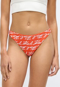 Červené plavky s bílým textem v kurzívě, nízký střih, hladký materiál a minimální boční pokrytí. Navrženo pro pou�žití jako plavky.