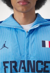 Helles blaues Sporttrikot aus strukturiertem Material, mit schwarzem "FRANCE" Schriftzug, Jordan-Logo und einer Applikation der französischen Flagge.